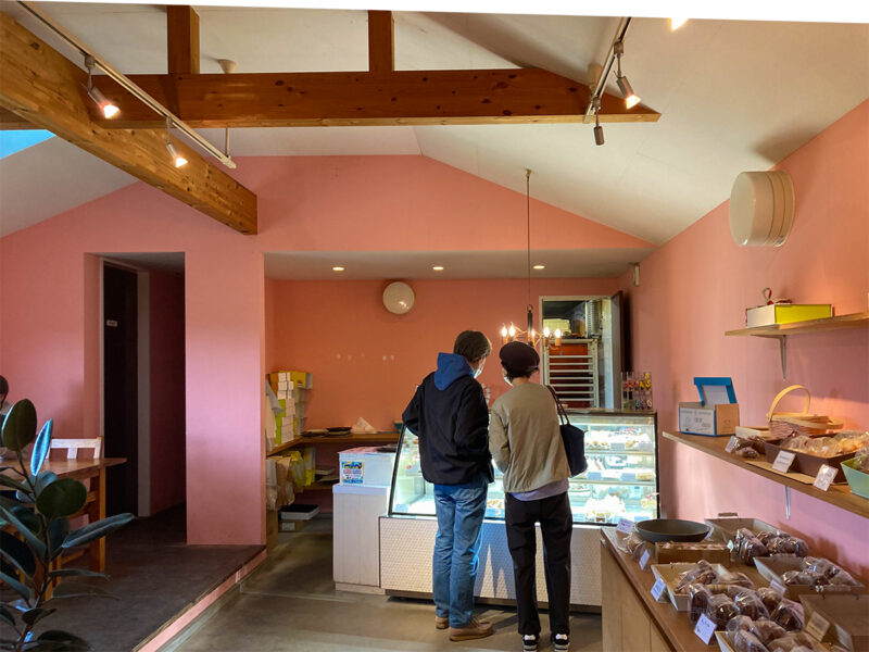 レジ側を見る「sweets & Café MILK」の内観。ピンク色の壁に囲まれ、両側にテーブル席と焼き菓子が並び、奥にはお手洗いと厨房があります